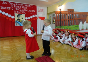 Na tle biało czerwonej dekoracji ubrane odświętnie dzieci recytują wiersz.